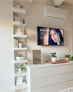 a flat screen tv mounted on the wall above a white shelf with plants and books