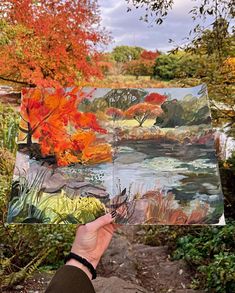 a person holding up an art piece in front of trees with orange leaves on it