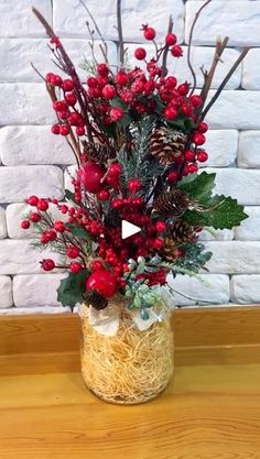 a vase filled with red berries and greenery on top of a wooden table next to a brick wall