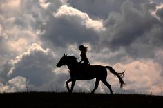 a woman riding on the back of a brown horse under cloudy skies with clouds in the background