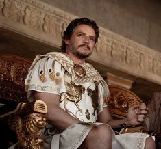 a man sitting on top of a wooden chair wearing a white shirt and gold trim