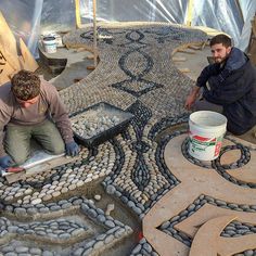 two men working on a mosaic design in a garden area with large rocks and gravel