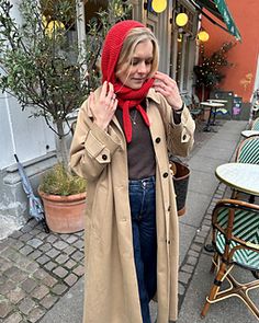 a woman in a trench coat and red scarf standing on the sidewalk with her hand up to her ear