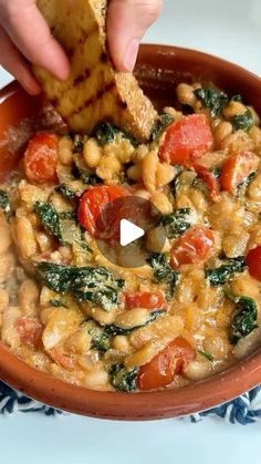 a person dipping some bread into a bowl of beans and spinach soup with tomatoes