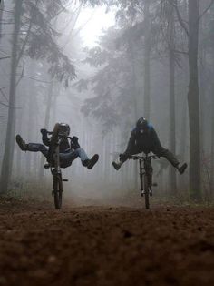 two people riding bikes in the woods on a foggy day with their feet up