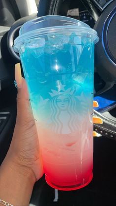 a hand holding up a cup with blue and red liquid in it on the dashboard of a car