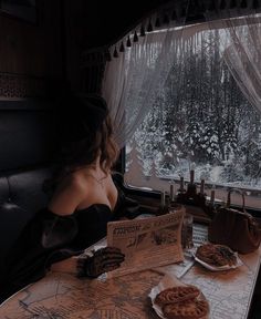 a woman sitting at a table looking out the window while reading a newspaper and eating cookies