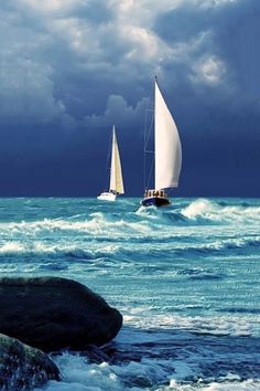 two sailboats sailing in the ocean on a cloudy day with rocks and crashing waves