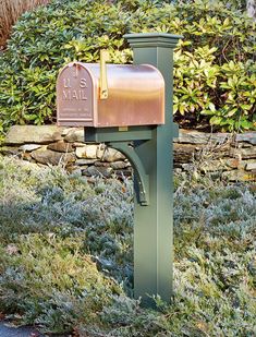 a green mailbox sitting in the grass next to a stone wall and shrubbery