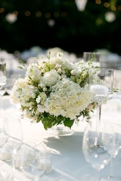 white flowers and greenery are arranged in a vase on a table with wine glasses