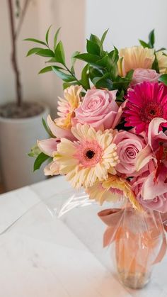 a vase filled with pink and yellow flowers on top of a white table next to a potted plant