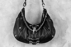 An ode to the timeless partnership of leather and chains, this leveled up black hobo bag is made from soft, premium genuine black leather and features a top shelf arrangement of silver hardware throughout.A centered, weighty metal industrial clip takes center stage, accompanied by an array of high contrast chromed out nailed rivets, centered feed through bars, and a draped top chain that doubles as a shoulder strap.Complete with a removable/adjustable leather and chain strap that can easily convert this purse from a shoulder bag to a comfortable crossbody bag, this design is an ode to the tough and timeless partnership of leather and chains.Bag is approx 13.5" x 7"Removable strap can extend up to 40"