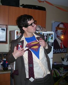 a man wearing a superman shirt and tie with a drink in his hand while standing next to a refrigerator