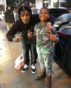 two young boys standing next to each other near a parked car and another person pointing at the camera