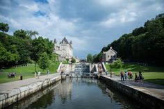 people are walking along the water in front of a large castle like building with stairs leading up to it