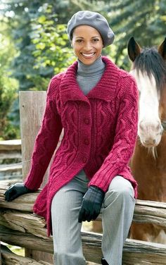 a woman sitting on a fence next to a horse wearing a red sweater and hat