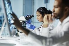 two people in lab coats looking through microscopes at something on a computer screen,