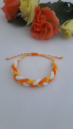 an orange and white rope bracelet next to some flowers on a white surface with one flower in the background