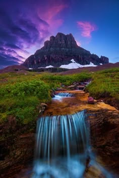 a small waterfall flowing into a lush green field under a purple sky with mountains in the background