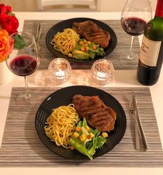 two black plates topped with meat and pasta next to glasses of wine on a table