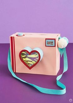 a pink box with a heart on it and a blue ribbon around the handle is sitting on a purple surface