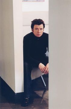 a man sitting on top of a white chair next to a wall and looking at the camera