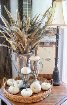 a table topped with a metal pitcher filled with white pumpkins and grass next to a lamp