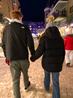 two people holding hands walking down the street at night with buildings in the back ground