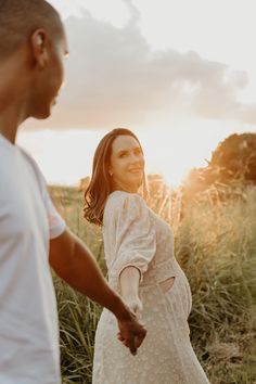 a pregnant woman holding the hand of her husband as they walk through tall grass at sunset