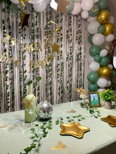 a table topped with balloons and confetti next to a wall covered in greenery