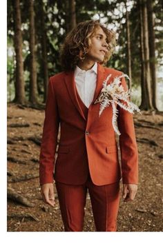 a man in an orange suit and white shirt standing in the woods with his hands in his pockets