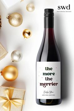 a bottle of wine sitting next to christmas ornaments and presents on a white surface with the words, she more the merrier