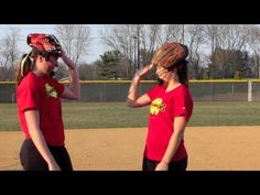 two girls in red shirts standing on a baseball field with their mitts over their heads