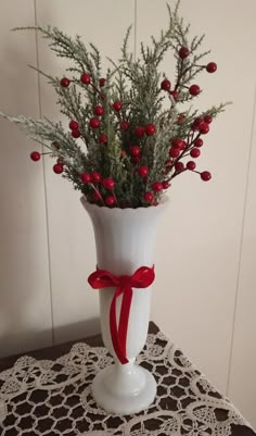 a white vase with red berries and greenery in it on a doily table