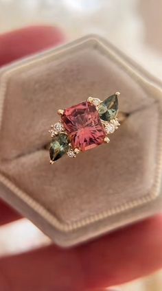 a pink ring with green and red stones in it's display case, sitting on top of a box