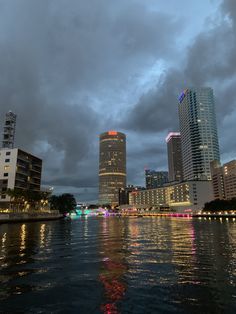 the city skyline is lit up at night with lights reflecting in the water and dark clouds