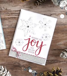 two christmas cards sitting on top of a wooden table next to pine cones and snowflakes