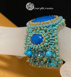 This is not the matching bracelet to my Mermaid's Attire set. It is very similar except light gold beads accent it, instead of the bronze colored beads in the full set. It features a large dyed howlite stone surrounded by Swarovski crystals. The center, with its embellishments measures 2.25 inches. The sides measure 2 inches. An aluminum cuff supports the bracelet and it fits mosts wrists. High wear ultrasuede lines the back. Based on a design by Sherry Serafini. Elegant Beaded Cuff Bracelets, Wearable Art Beaded Cuff Bracelet Gift, Blue Beaded Cuff Bracelet Gift, Sherry Serafini, Luxury Beaded Statement Cuff Bracelet, Turquoise Hand-strung Stretch Bracelet For Beach, Howlite Stone, Turquoise Cuff, Matching Bracelet