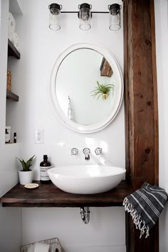 a white sink sitting under a round mirror on top of a wooden shelf next to a potted plant