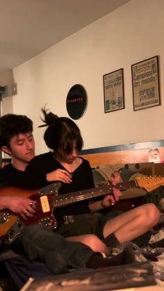 two people sitting on a bed with guitars in their hands and one playing the guitar