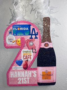 a pink and glitter number two sign with a bottle of champagne next to it on a white wall