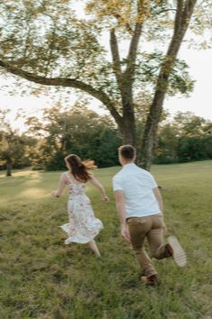 a man and woman are running through the grass in front of a tree with their arms around each other