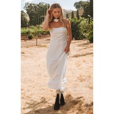 a woman wearing a white dress and black booties standing in the dirt with her hand on her head