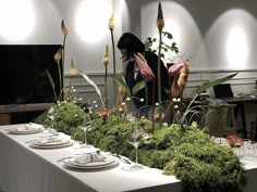 a woman standing next to a long table covered in flowers and greenery with plates on it