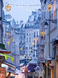 people are walking down an alley way with lights strung from the buildings in the background