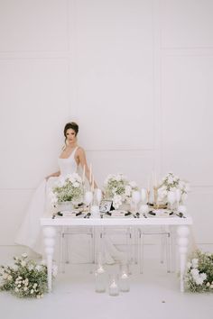 a woman sitting at a table with white flowers and candles on it, surrounded by greenery