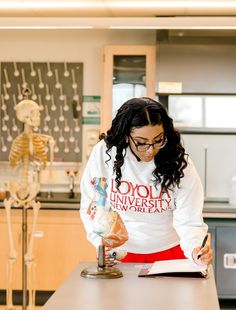 a woman in a white sweatshirt is looking at a book on a table with skeleton models behind her