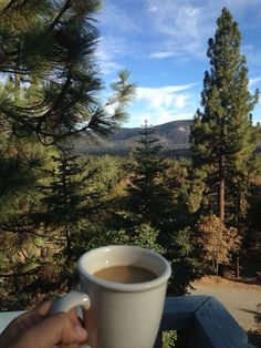 a person holding a cup of coffee on top of a wooden table in front of some trees