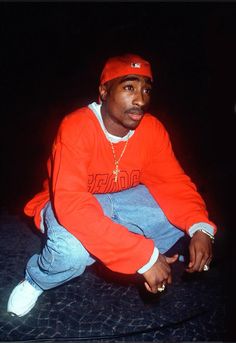 a man sitting on the ground wearing an orange sweatshirt and jeans with his hands in his pockets