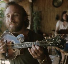 a man holding a guitar in his right hand and looking at the camera while people sit around him